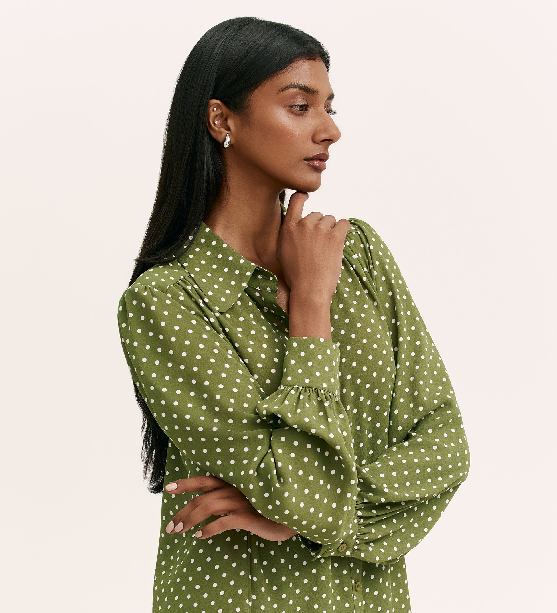 A woman with long dark hair wears the Melina Green Spot Chiffon Blouse—an olive green relaxed fit top with white polka dots. She stands against a light background, looking to the side, thoughtfully touching her chin.