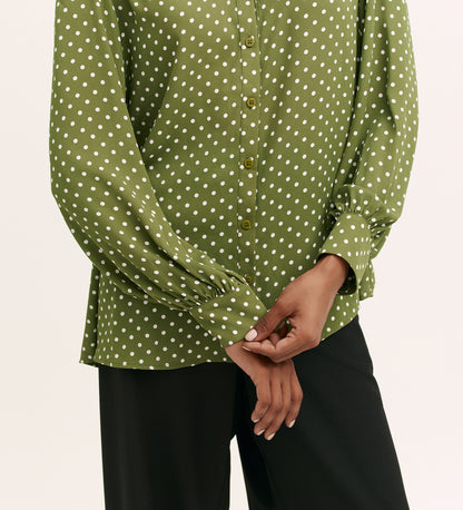 A person wears the Melina Green Spot Chiffon Blouse with black pants. Their hands are loosely clasped in front, and only their torso and arms are visible.