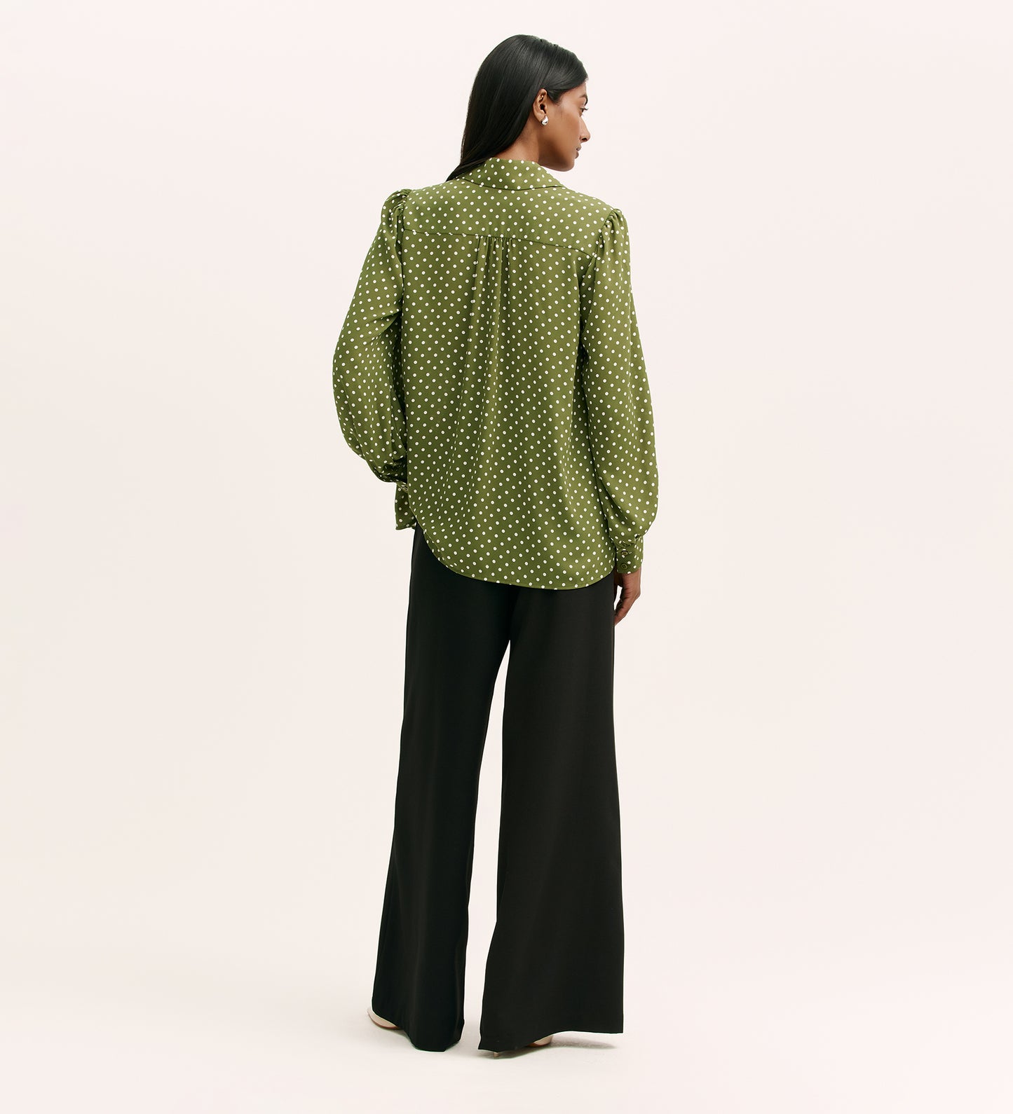 A woman stands facing away, wearing the Melina Green Spot Chiffon Blouse with black wide-leg pants against a plain light background.