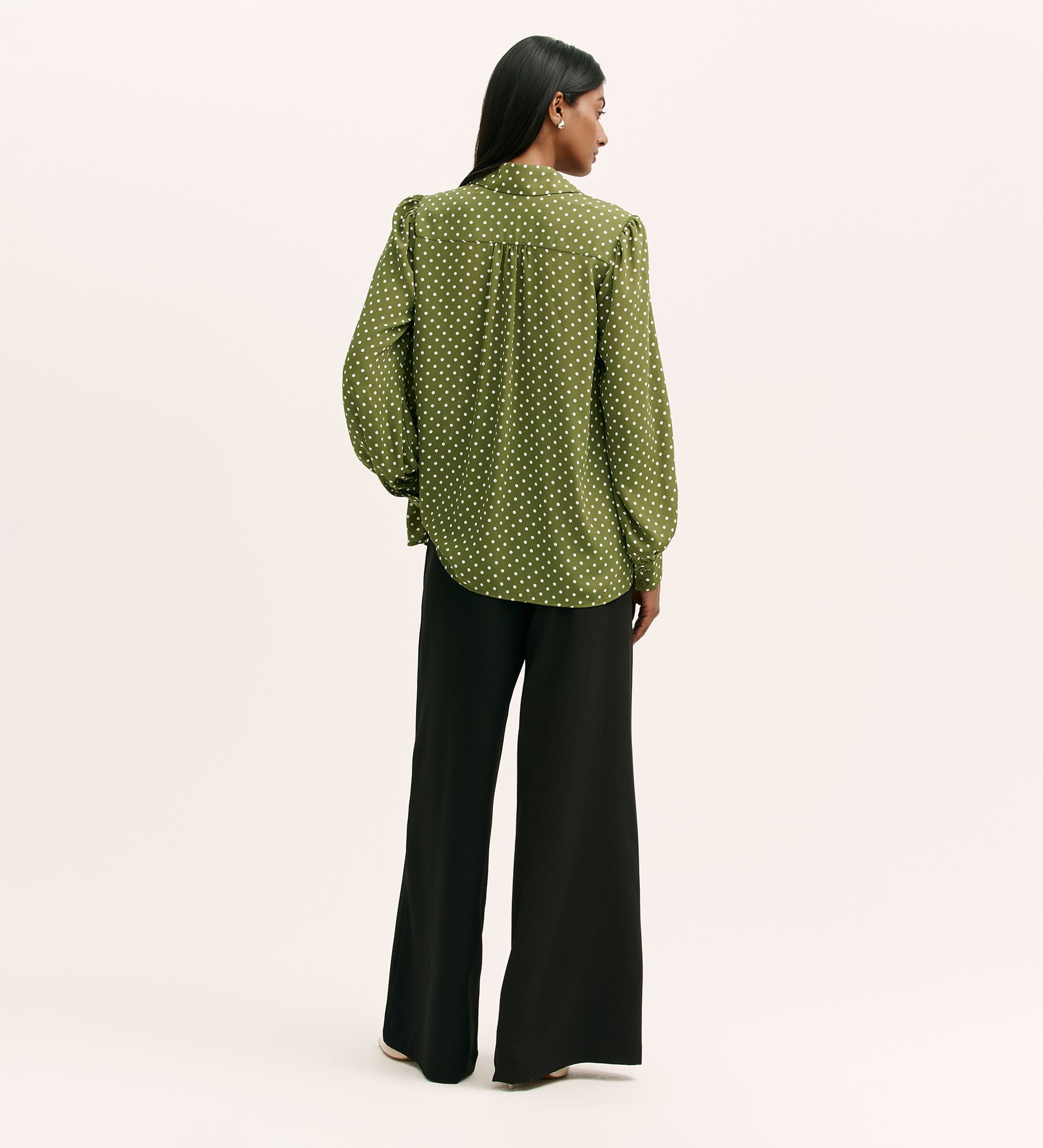 A woman stands facing away, wearing the Melina Green Spot Chiffon Blouse with black wide-leg pants against a plain light background.