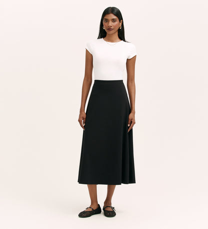 A woman stands against a plain background wearing a fitted white short-sleeve top, the Ruth Black Ponte Jersey Midi Skirt with an elasticated waistband, and black flat shoes. Her hair is straight and she wears minimal accessories.