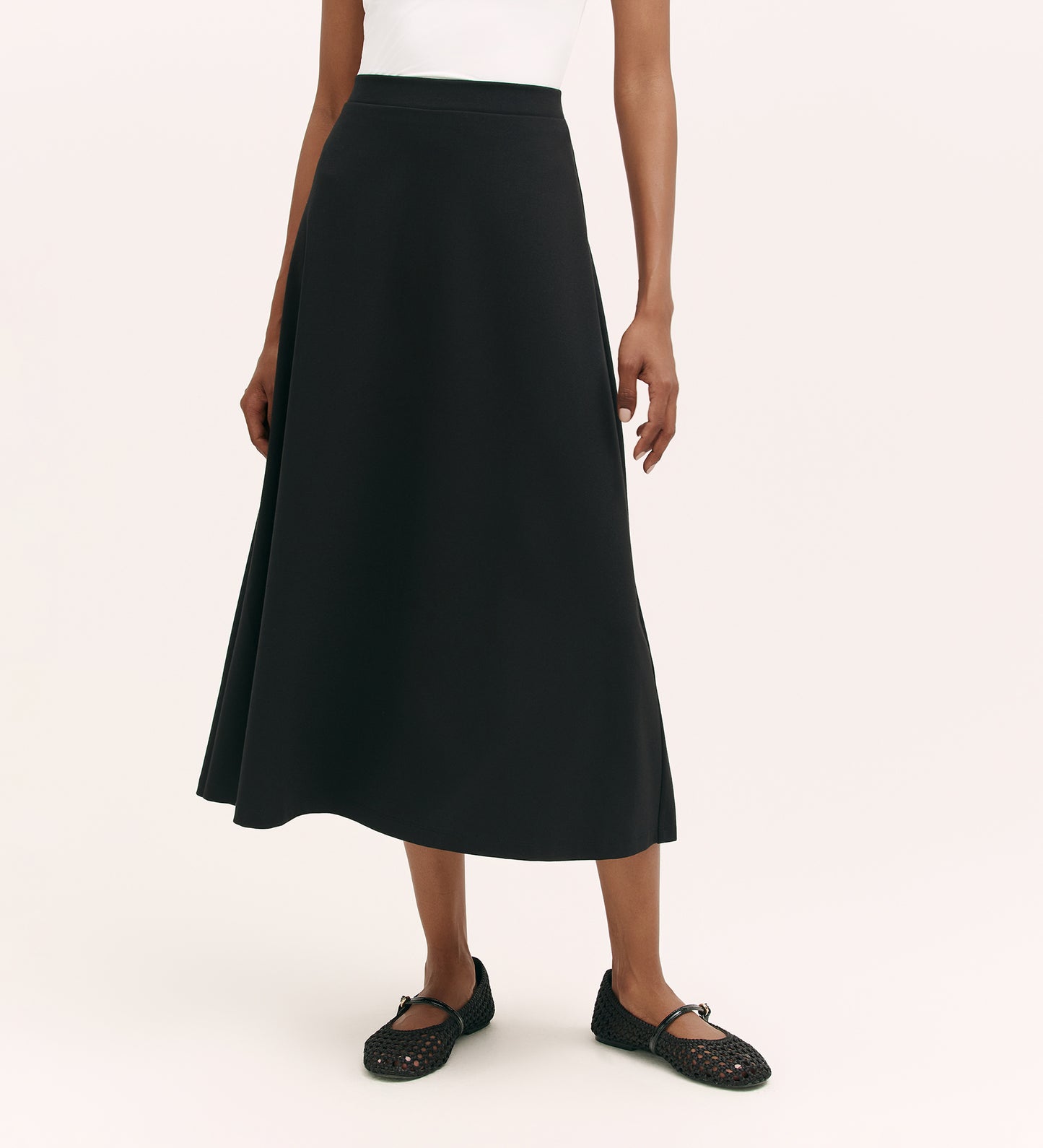 A person in a white sleeveless top and the Ruth Black Ponte Jersey Midi Skirt with an elasticated waistband, paired with black woven flats, stands against a plain light background.