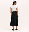A woman with straight dark hair faces away, wearing a short-sleeved white top and the Ruth Black Ponte Jersey Midi Skirt, which is high-waisted with an elastic waistband. She pairs it with black flats against a plain light background.