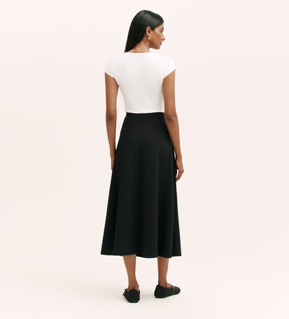 A woman with straight dark hair faces away, wearing a short-sleeved white top and the Ruth Black Ponte Jersey Midi Skirt, which is high-waisted with an elastic waistband. She pairs it with black flats against a plain light background.