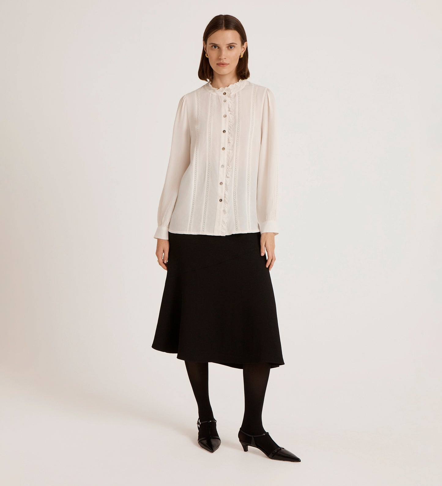 A woman with shoulder-length brown hair and a neutral expression stands against a plain background wearing the Flora Ivory Crepe Blouse, a black knee-length skirt, black tights, and pointed-toe shoes.