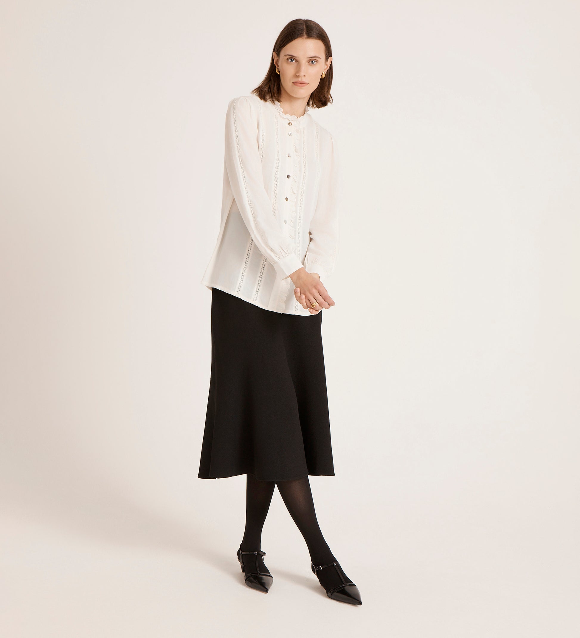 A woman with straight brown hair and a neutral expression stands against a plain background wearing the Flora Ivory Crepe Blouse, a black midi skirt, black tights, and pointed black shoes.