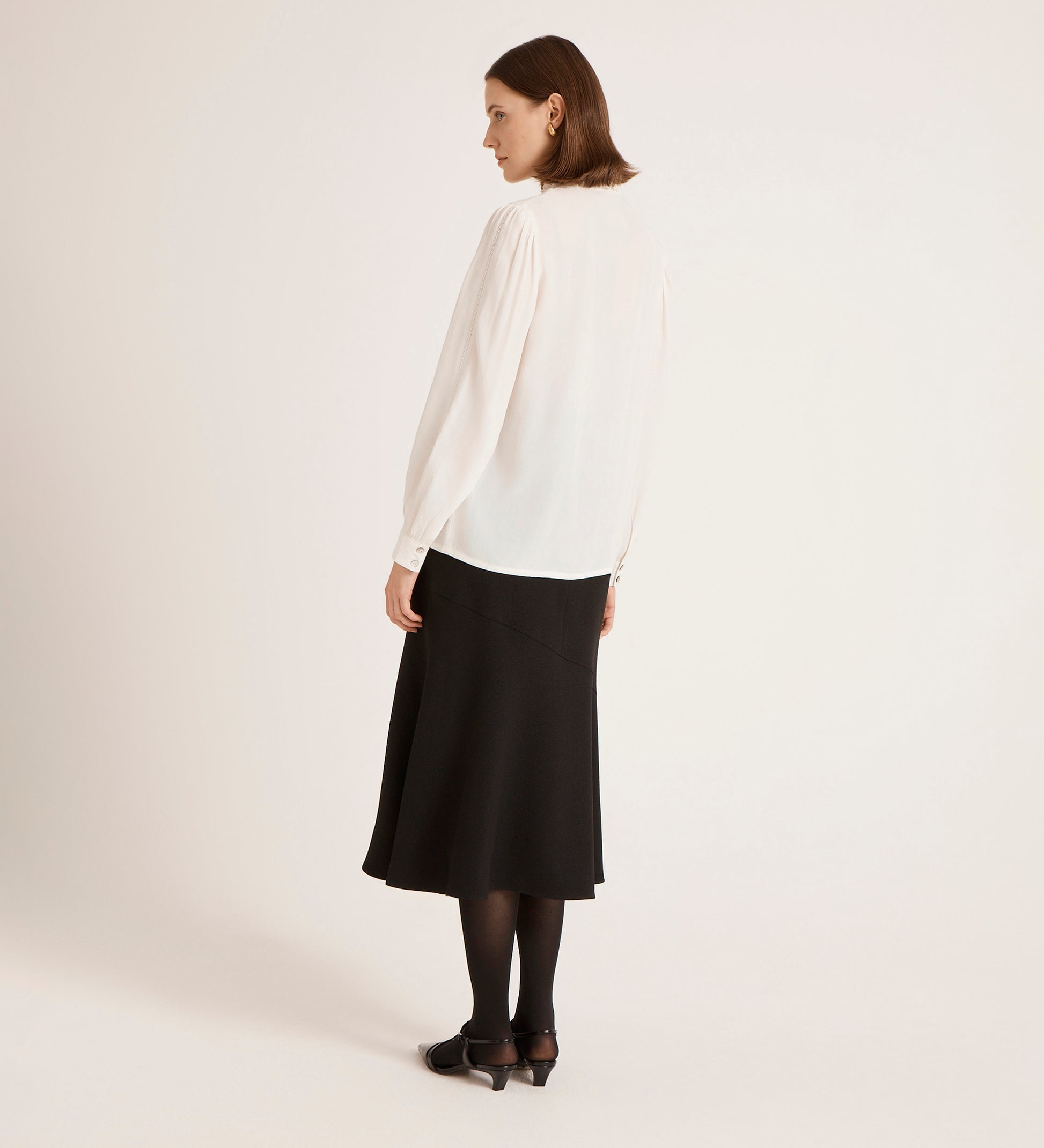 A woman, back turned, wears the Flora Ivory Crepe Blouse with a black midi skirt, tights, and shoes against a plain light background.