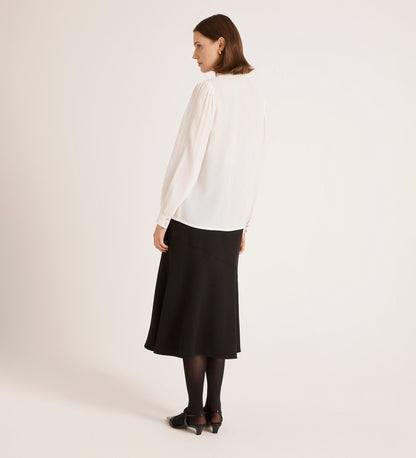 A woman, back turned, wears the Flora Ivory Crepe Blouse with a black midi skirt, tights, and shoes against a plain light background.