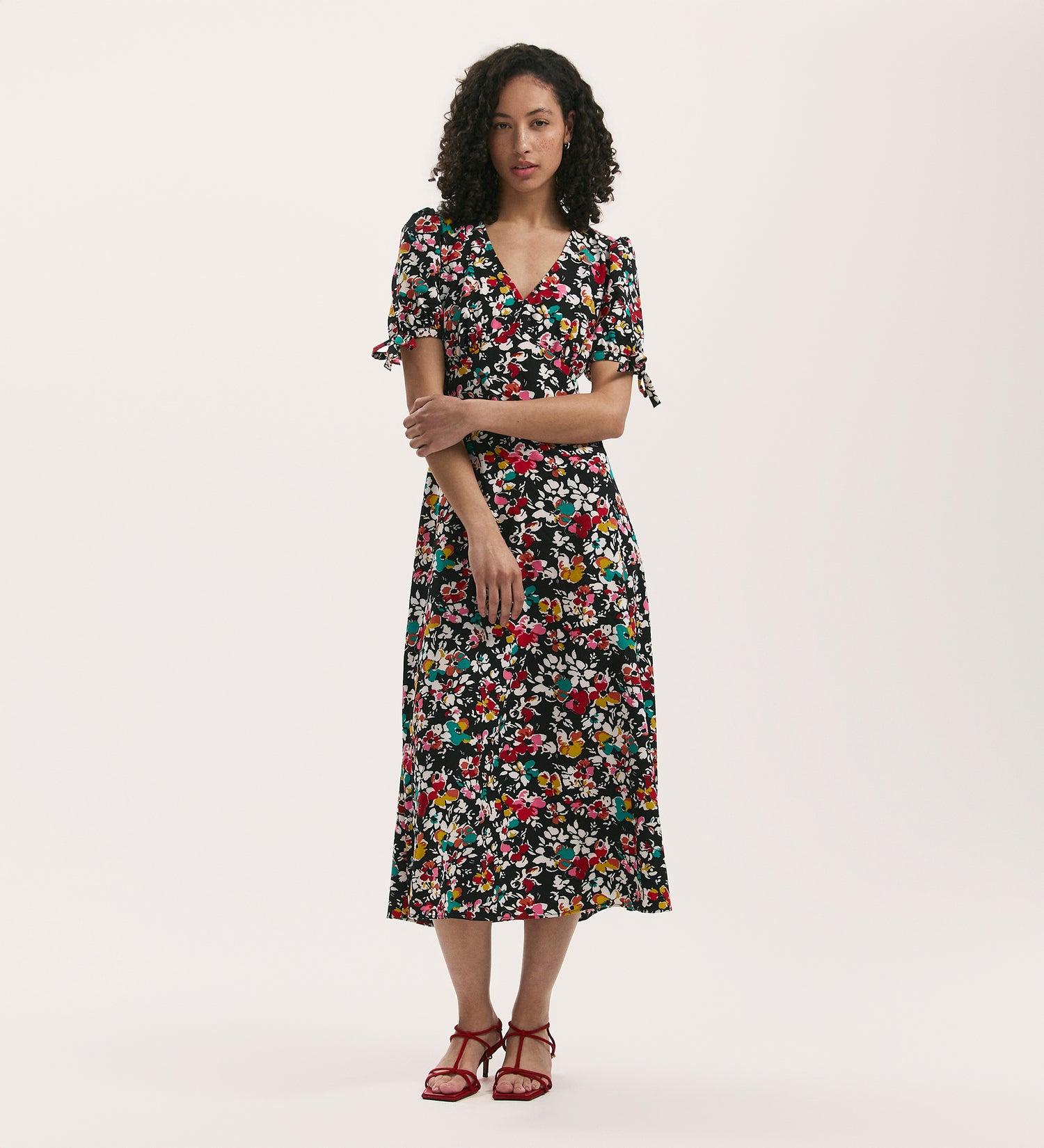 A woman with curly hair stands against a plain background in the Gabriella Black Floral Crepe Midi Tea Dress, made from crease-free crepe, featuring short sleeves. She pairs it with red strappy sandals and crosses her arms in front of her.