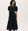 A woman with curly hair models the Genevive Black Cotton Midi Tiered Dress, which features puff sleeves and a tiered skirt, standing against a plain light background.