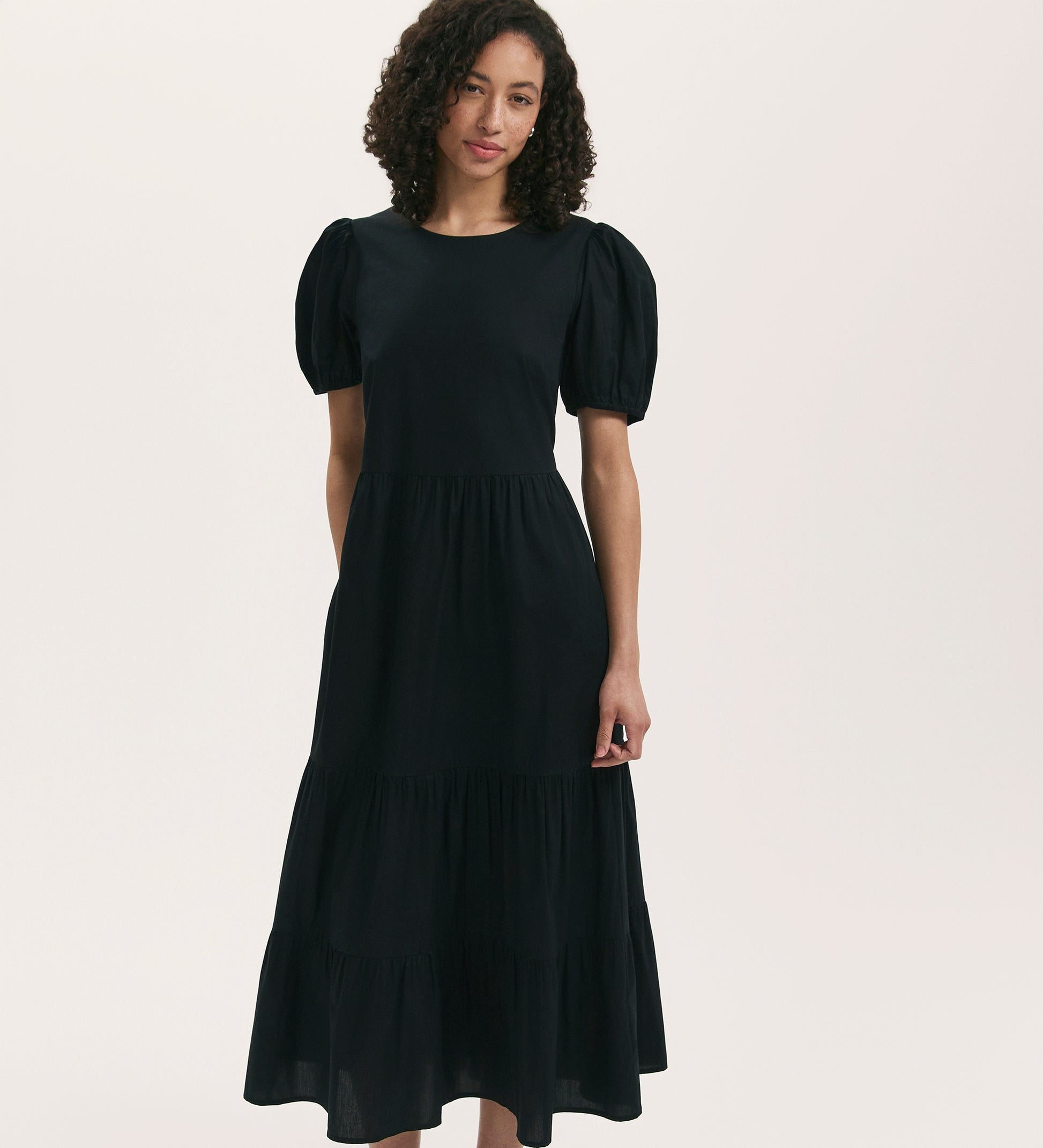 A woman with curly hair models the Genevive Black Cotton Midi Tiered Dress, which features puff sleeves and a tiered skirt, standing against a plain light background.