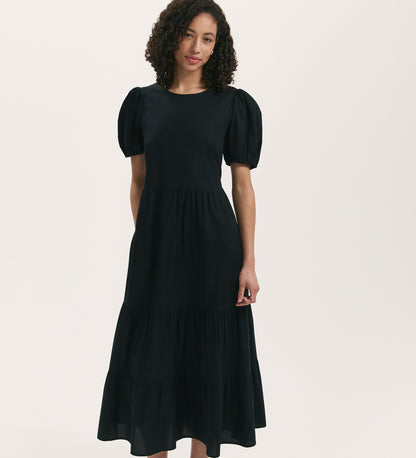 A woman with curly hair models the Genevive Black Cotton Midi Tiered Dress, which features puff sleeves and a tiered skirt, standing against a plain light background.