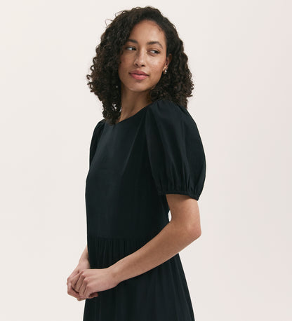 A woman with curly hair wears the Genevive Black Cotton Midi Tiered Dress with short puff sleeves, standing against a plain light background and looking to the side with a gentle expression.
