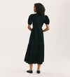 A woman with curly hair stands facing away, wearing the Genevive Black Cotton Midi Tiered Dress with puffed sleeves and black flats, set against a plain light background.