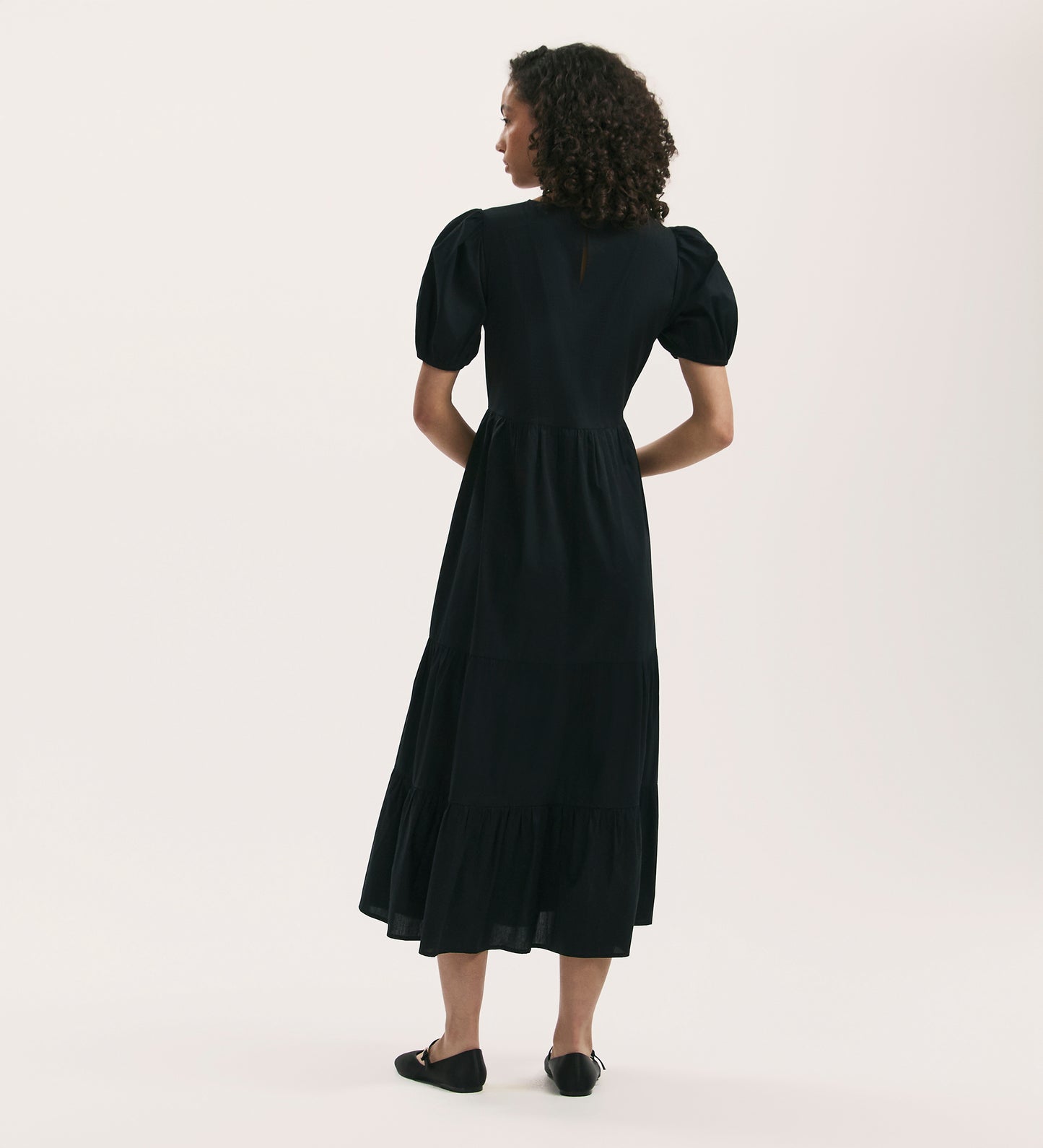 A woman with curly hair stands facing away, wearing the Genevive Black Cotton Midi Tiered Dress with puffed sleeves and black flats, set against a plain light background.