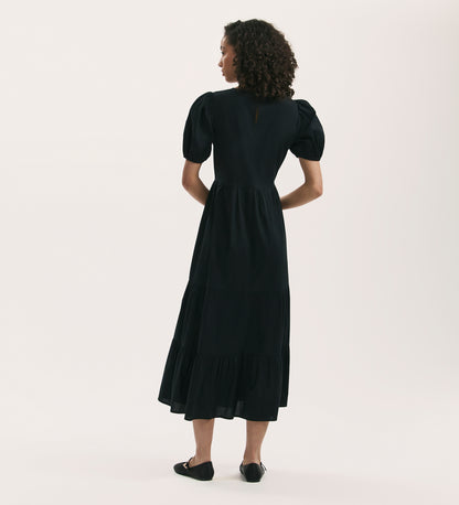 A woman with curly hair stands facing away, wearing the Genevive Black Cotton Midi Tiered Dress with puffed sleeves and black flats, set against a plain light background.