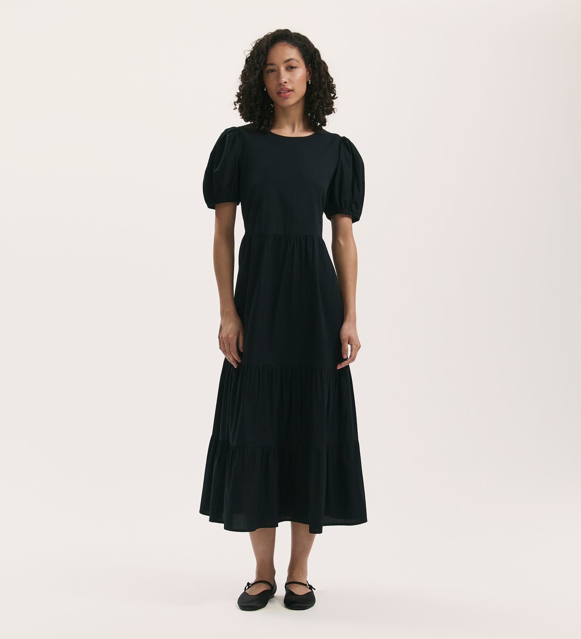 A woman with curly hair stands against a plain light background wearing the Genevive Black Cotton Midi Tiered Dress, featuring short sleeves, paired with black flats.