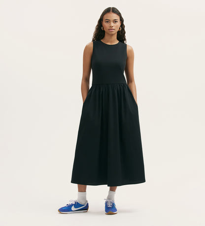 A woman with long curly hair wears the Georgie Black Cotton Sleeveless Midi Dress, paired with white socks and blue Nike sneakers, standing against a plain light background.