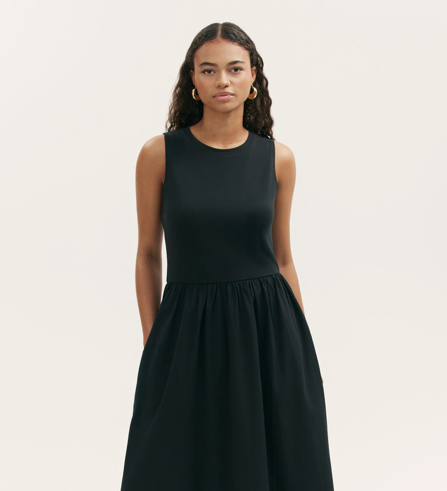 A woman with long, curly hair stands against a light background, wearing the Georgie Black Cotton Sleeveless Midi Dress with a fitted bodice and relaxed fit, hands in her pockets and a neutral expression.