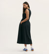 A woman stands sideways, looking over her shoulder, wearing the Georgie Black Cotton Sleeveless Midi Dress with pockets and a relaxed fit, paired with white socks and blue sneakers against a plain light background.