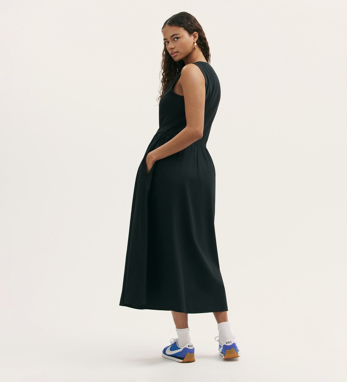 A woman stands sideways, looking over her shoulder, wearing the Georgie Black Cotton Sleeveless Midi Dress with pockets and a relaxed fit, paired with white socks and blue sneakers against a plain light background.