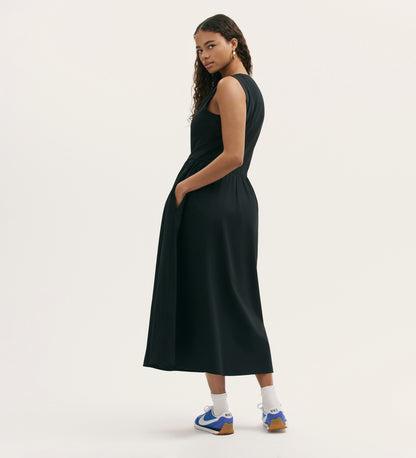 A woman stands sideways, looking over her shoulder, wearing the Georgie Black Cotton Sleeveless Midi Dress with pockets and a relaxed fit, paired with white socks and blue sneakers against a plain light background.