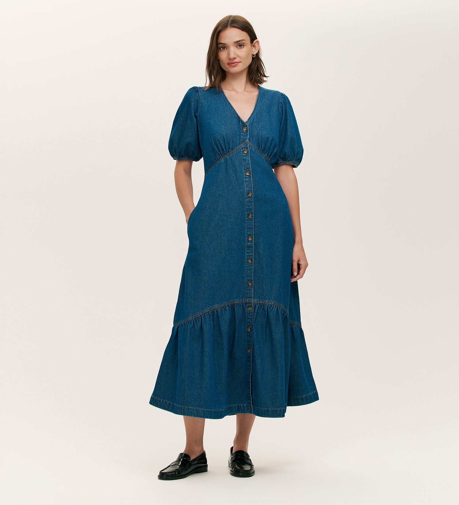 A woman faces forward wearing the Goldie Mid Blue Denim Midi Dress, featuring short puff sleeves, a V-neckline, and button-through front. She pairs it with black shoes against a plain light background.