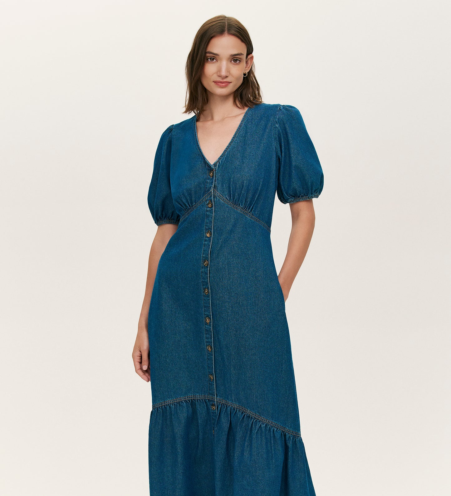 A woman with straight brown hair models the Goldie Mid Blue Denim Midi Dress, featuring puffed short sleeves, a V-neckline, button-through front, and a tiered skirt, standing against a plain light background.