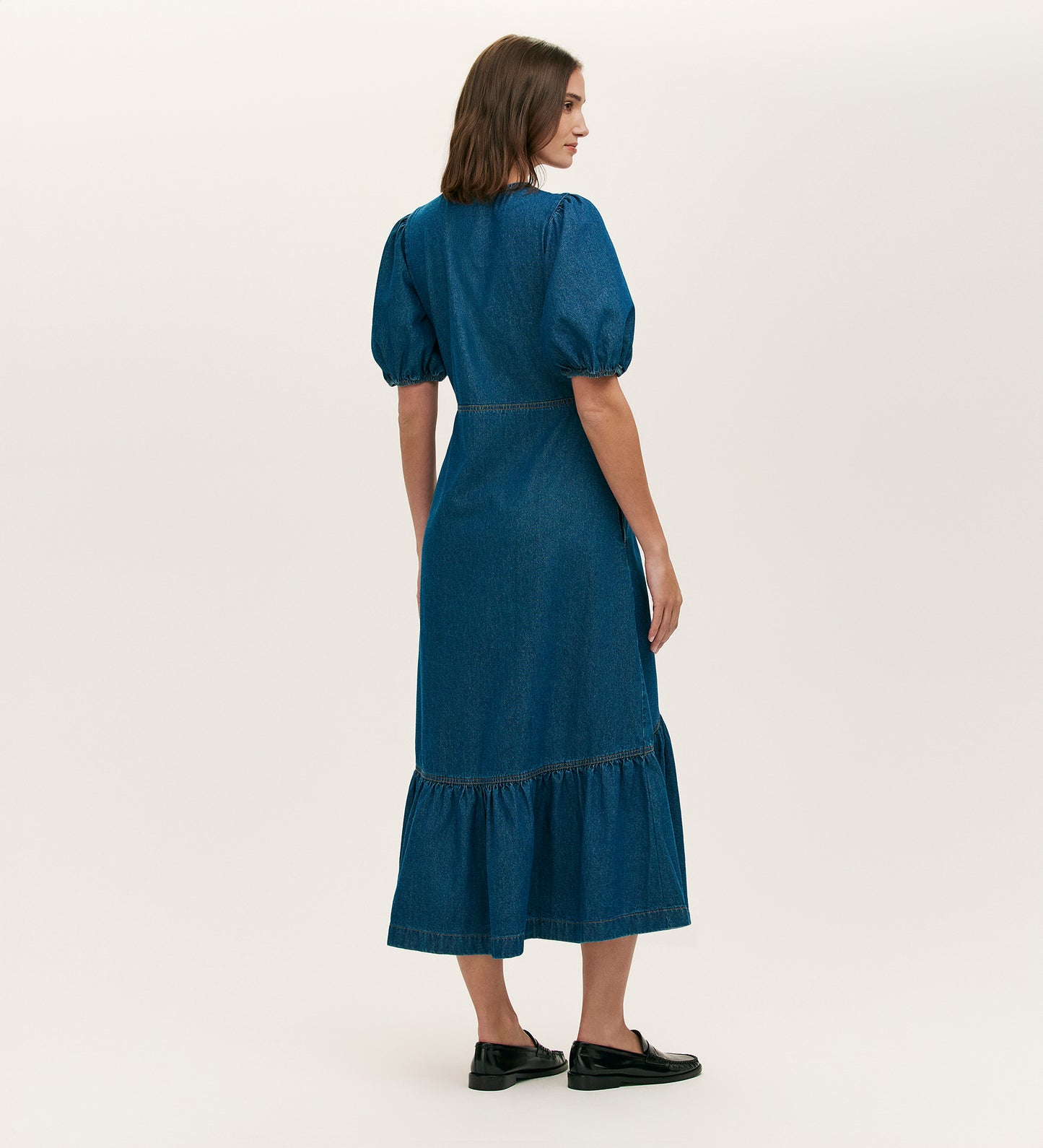 A woman with shoulder-length brown hair wears the Goldie Mid Blue Denim Midi Dress, featuring puffed short sleeves and a tiered skirt. She stands facing away in black flats against a plain light background.