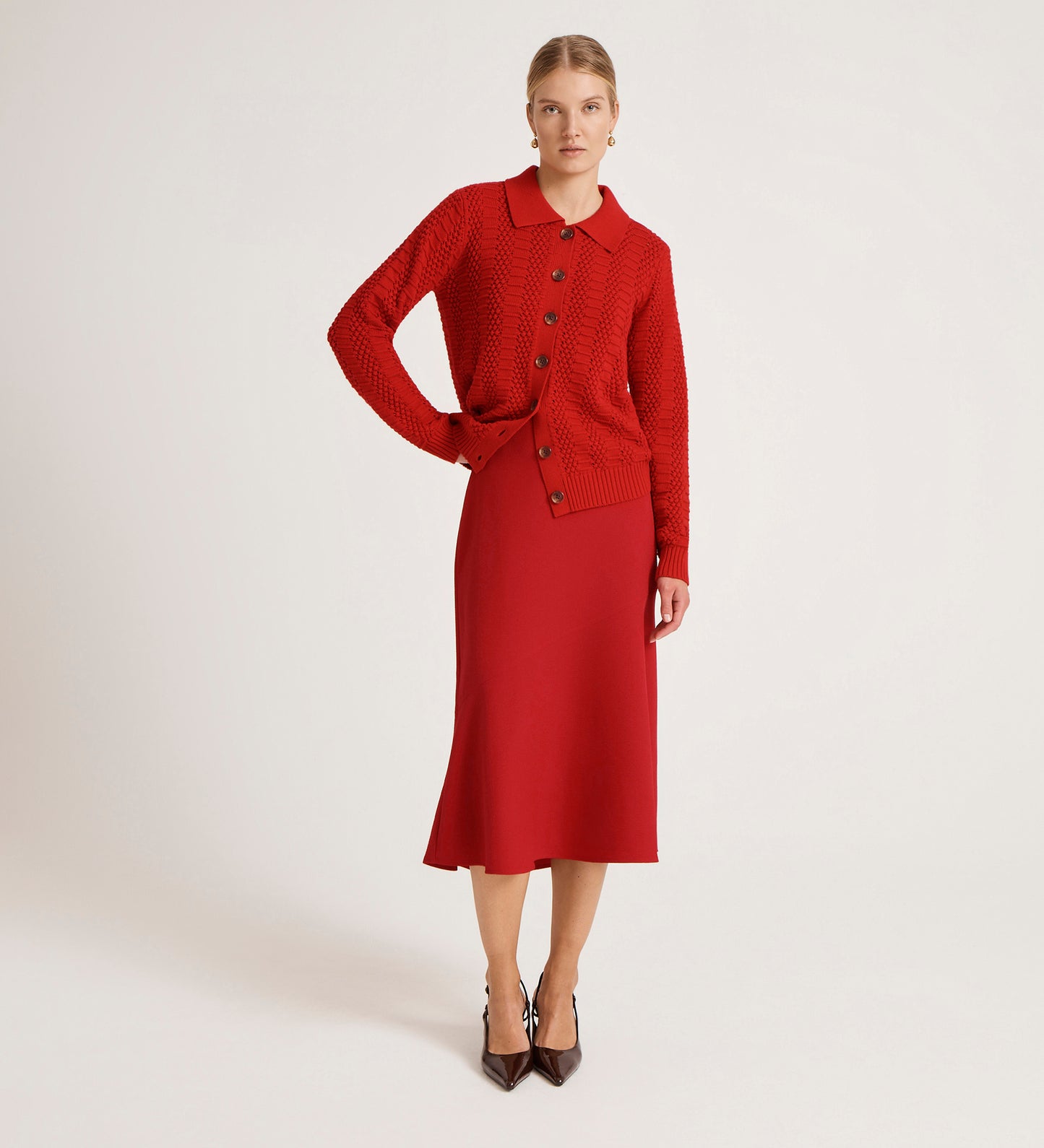 A woman stands against a plain background wearing a red button-up knit cardigan and the Gracie Red Rich Crepe Midi Skirt with a flared silhouette, paired with brown pointed-toe heels.