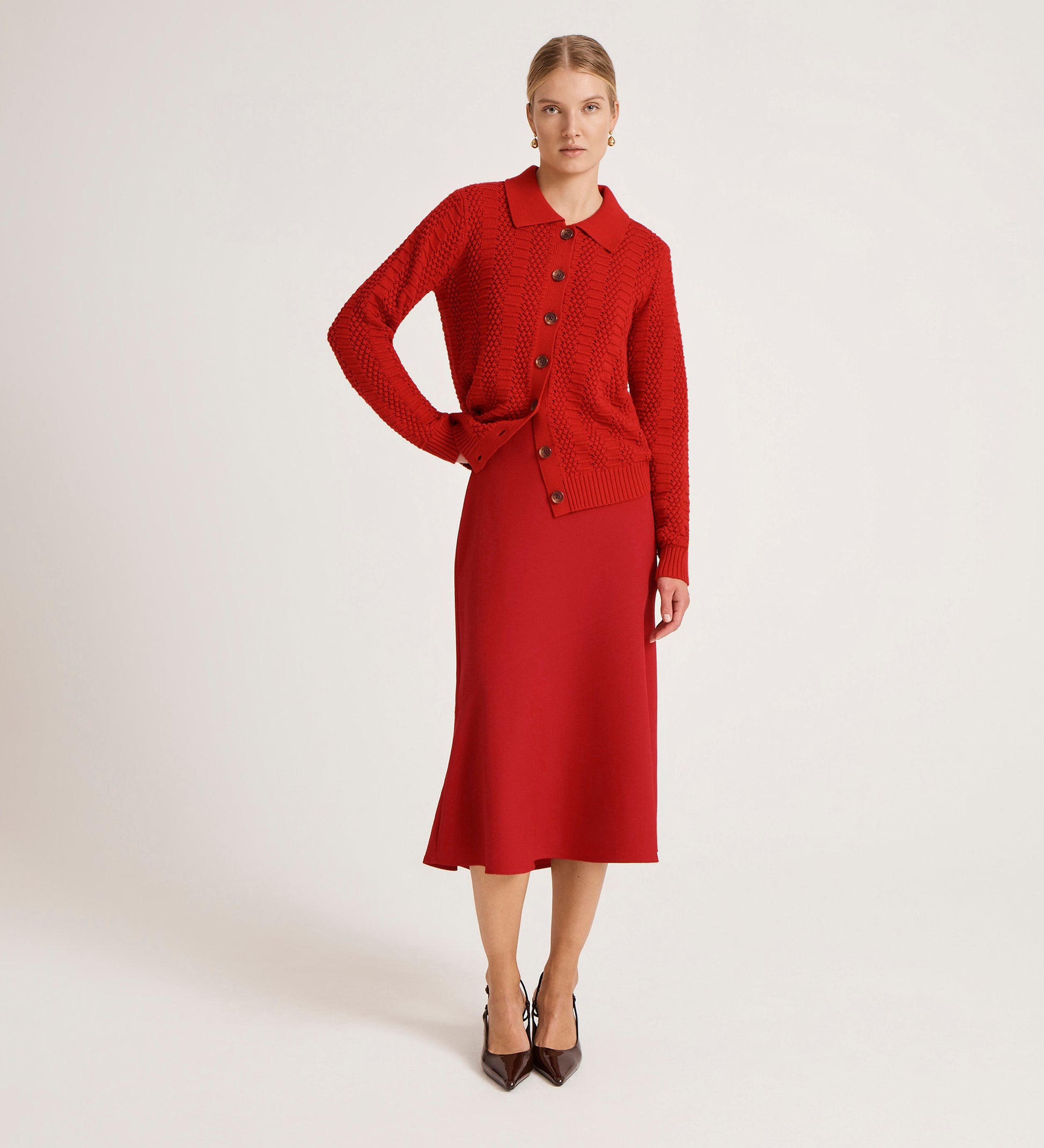A woman stands against a plain background wearing a red button-up knit cardigan and the Gracie Red Rich Crepe Midi Skirt with a flared silhouette, paired with brown pointed-toe heels.