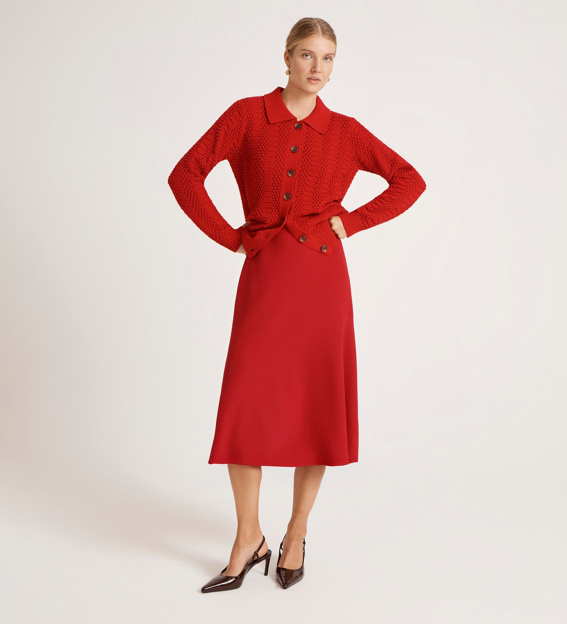 A woman stands against a plain background wearing the Gracie Red Rich Crepe Midi Skirt and a matching red sweater, paired with dark pointed-toe heels. Her blonde hair is pulled back, and she rests her hands on her hips.