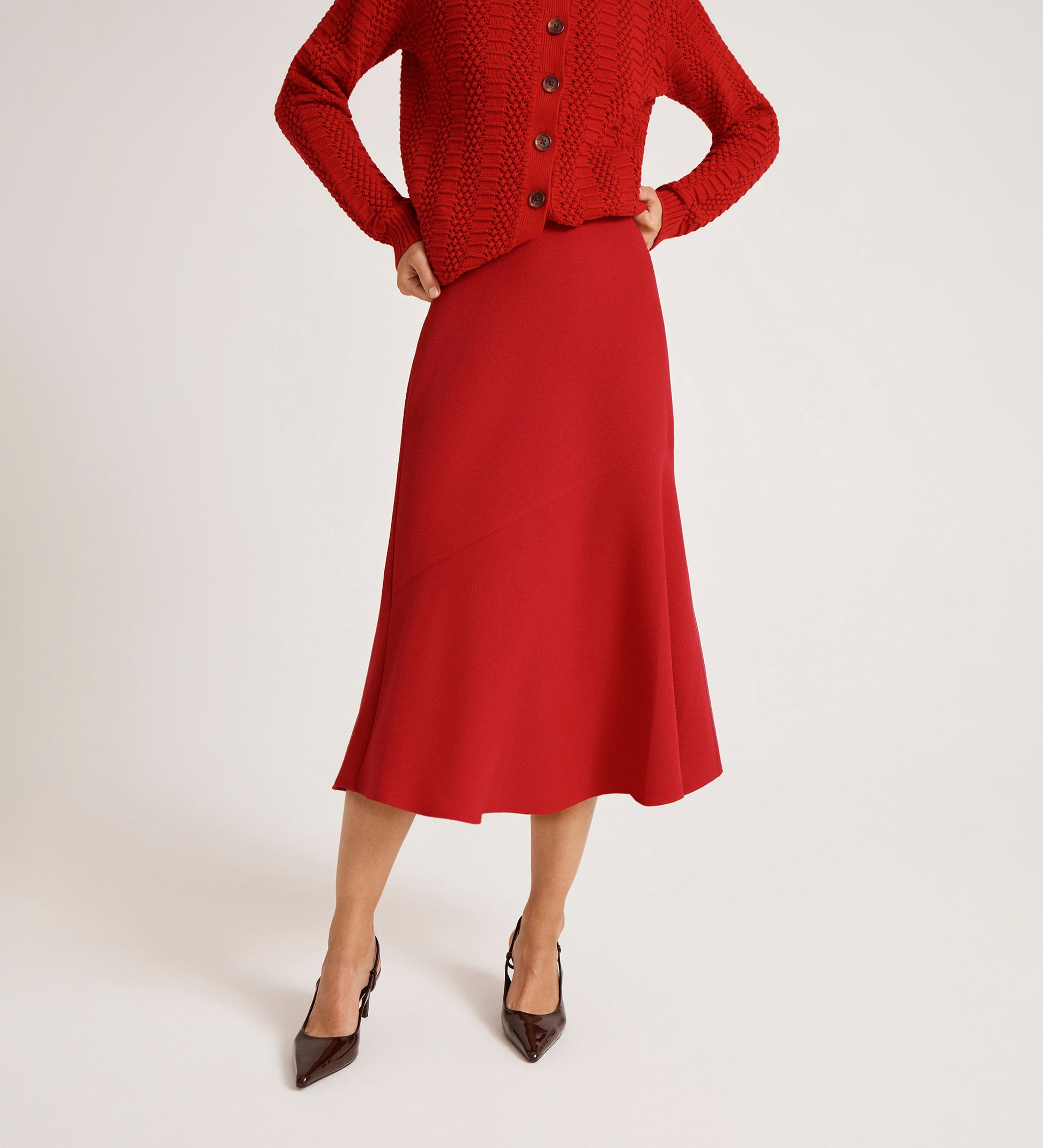 A person wears the Gracie Red Rich Crepe Midi Skirt and a matching red cardigan with a flared silhouette, paired with pointed brown heels, hands on hips, against a white background.