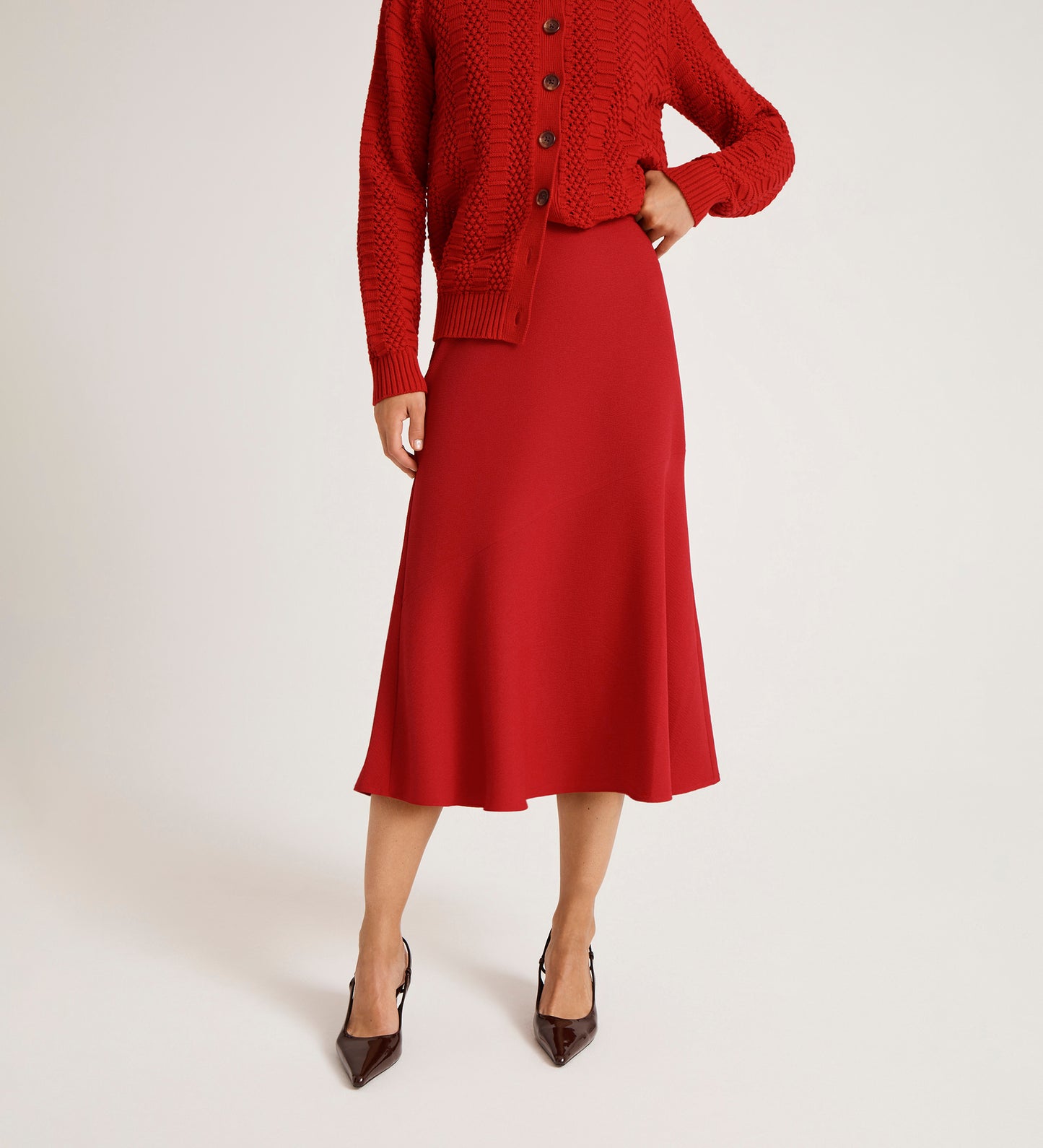 Wearing a red knit sweater and the Gracie Red Rich Crepe Midi Skirt, paired with black pointed-toe heels, a person poses against a plain white background with their face out of view.