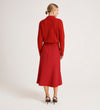 A woman with blonde hair in a low bun stands against a white background, wearing a red textured sweater, black heeled sandals, and the Gracie Red Rich Crepe Midi Skirt.
