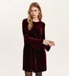 A woman with long blonde hair models the Hattie Burgundy Velvet Short Dress, featuring long flared sleeves. She stands against a plain light background, looking to the side with a neutral expression, embodying effortless party style.