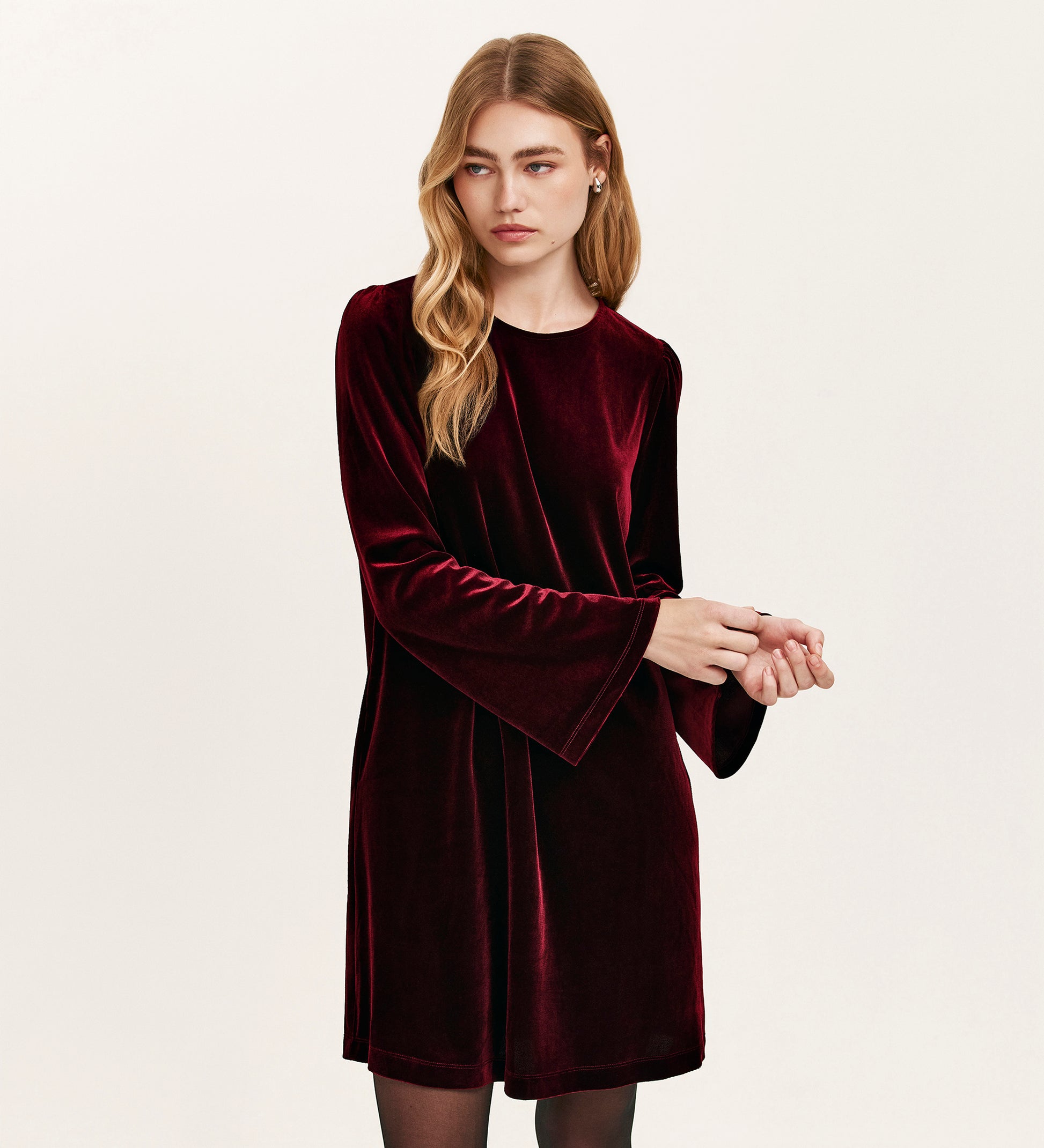 A woman with long blonde hair models the Hattie Burgundy Velvet Short Dress, featuring long flared sleeves. She stands against a plain light background, looking to the side with a neutral expression, embodying effortless party style.