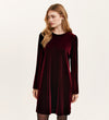A woman with long blonde hair wears the Hattie Burgundy Velvet Short Dress, a long-sleeved, knee-length jersey velvet piece—an ideal party outfit—while standing against a plain light background, looking slightly to her left.