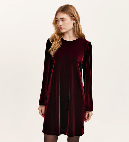 A woman with long blonde hair wears the Hattie Burgundy Velvet Short Dress, a long-sleeved, knee-length jersey velvet piece—an ideal party outfit—while standing against a plain light background, looking slightly to her left.