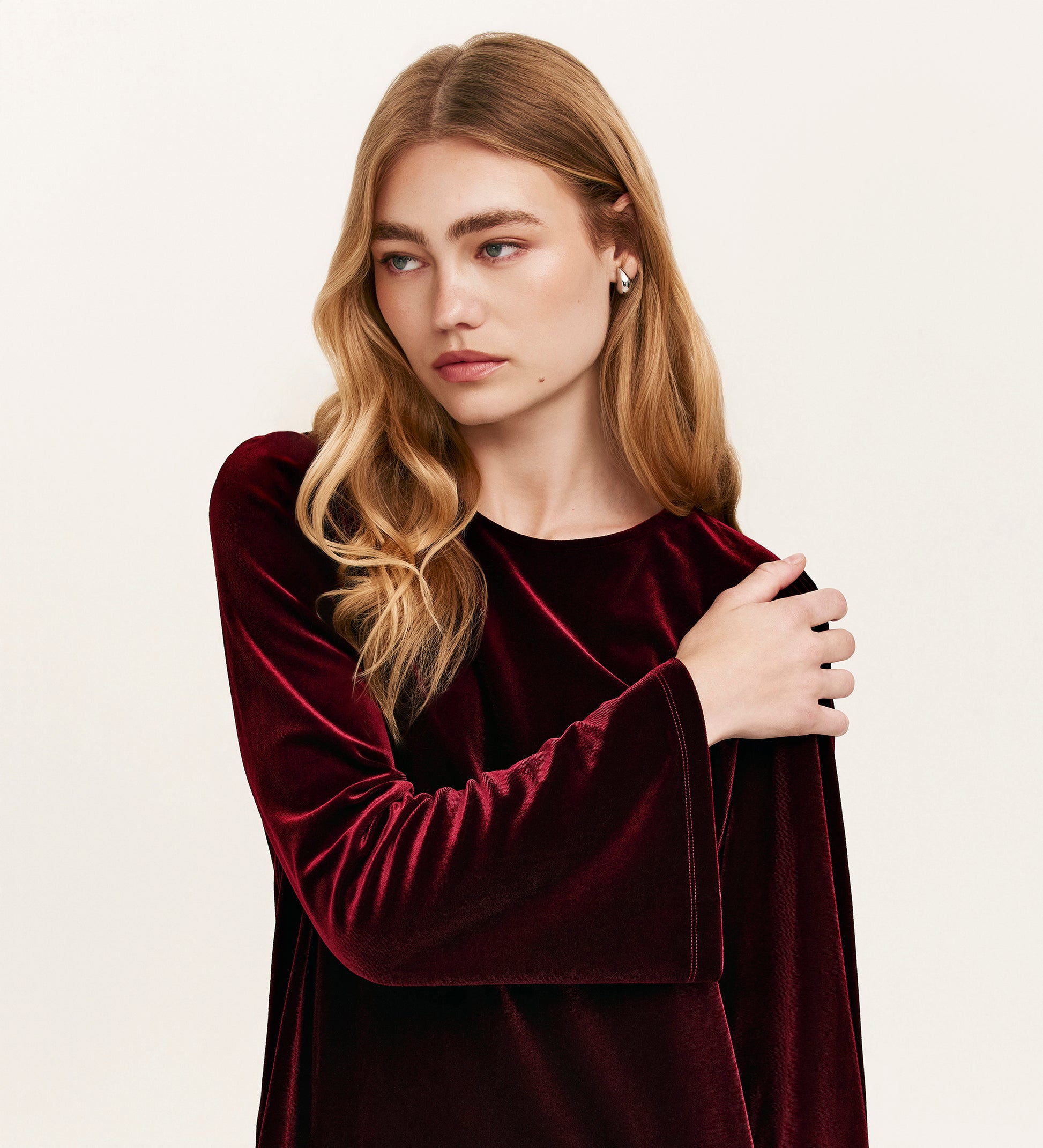 A young woman with long, wavy blonde hair models the Hattie Burgundy Velvet Short Dress. She looks left with a neutral expression, her left arm gently resting on her right shoulder, effortlessly embodying party style against a light background.