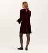 A woman faces away, wearing the Hattie Burgundy Velvet Short Dress with long sleeves—ideal for parties—paired with black tights and shoes. She stands against a plain, light-colored background.