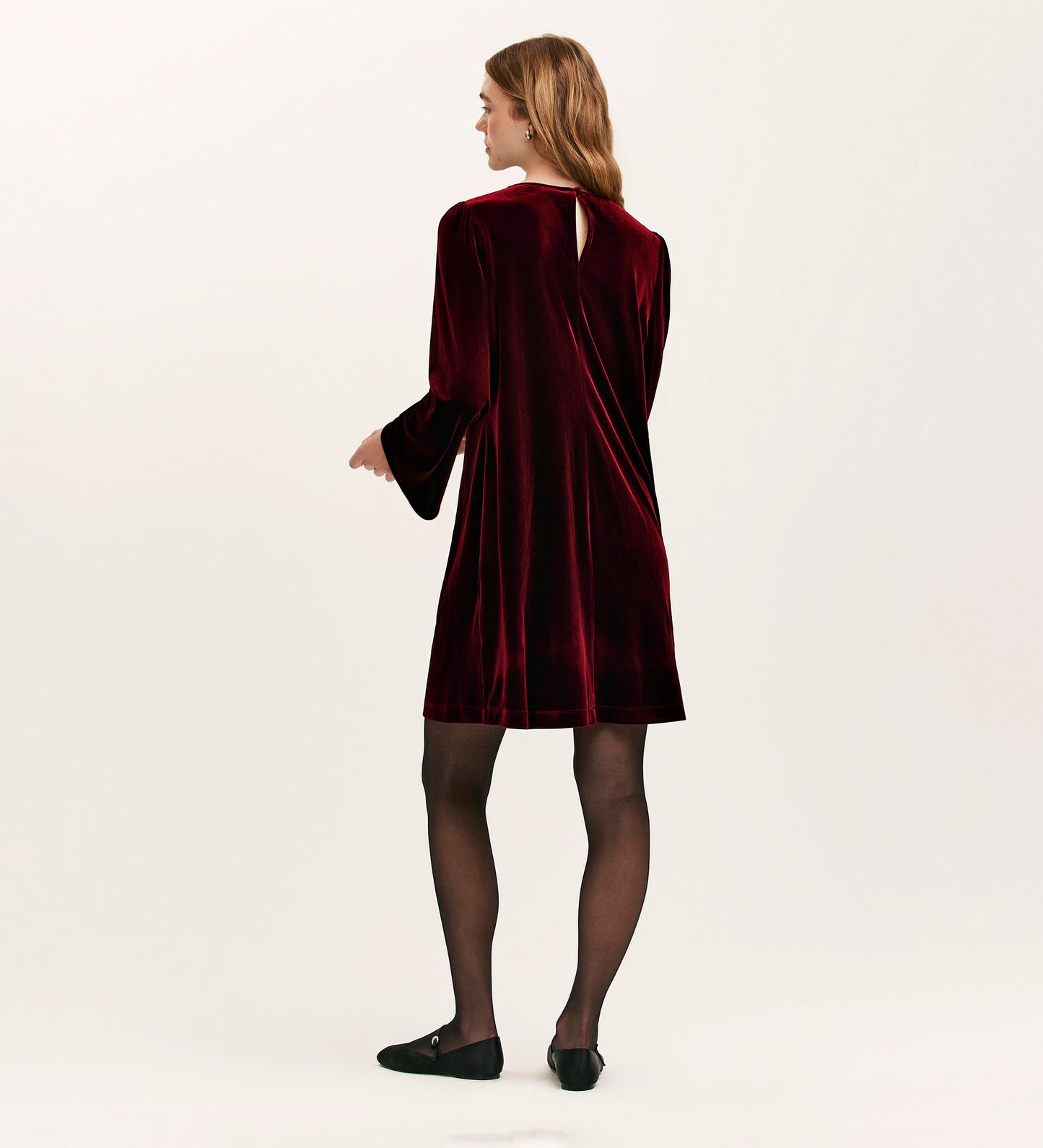A woman faces away, wearing the Hattie Burgundy Velvet Short Dress with long sleeves—ideal for parties—paired with black tights and shoes. She stands against a plain, light-colored background.