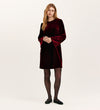A woman with long, wavy blonde hair wears the Hattie Burgundy Velvet Short Dress with bell sleeves, styled with black sheer tights and flat shoes—an ideal choice for effortless party dressing.