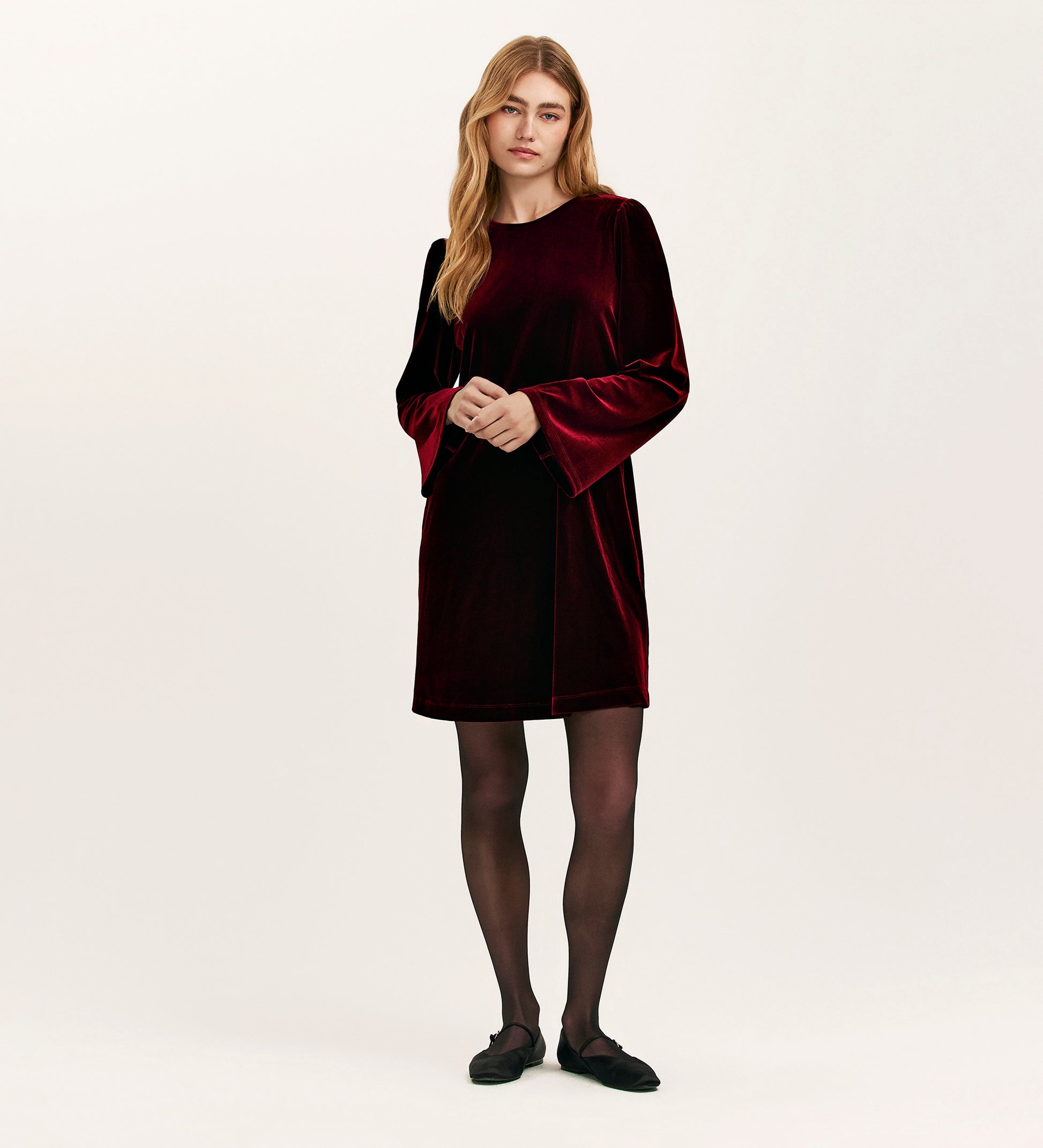 A woman with long, wavy blonde hair wears the Hattie Burgundy Velvet Short Dress with bell sleeves, styled with black sheer tights and flat shoes—an ideal choice for effortless party dressing.