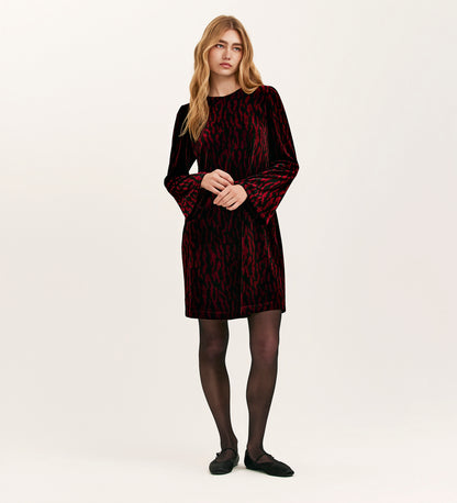 A woman with long blonde hair wears the Hattie Burgundy Zebra Jersey Velvet Short Dress, paired with black tights and black flats, standing against a plain light background.