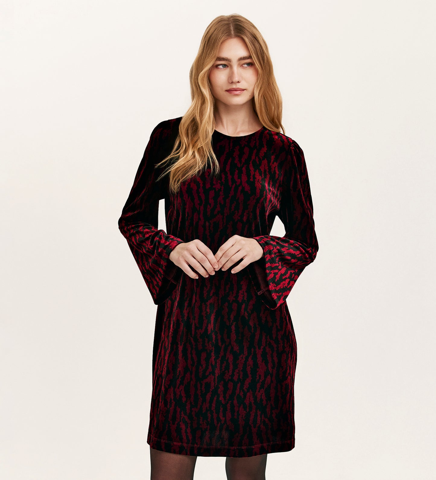 A woman with long blonde hair wears the Hattie Burgundy Zebra Jersey Velvet Short Dress, featuring a red animal print and bell sleeves, perfect for parties, standing against a plain light background.