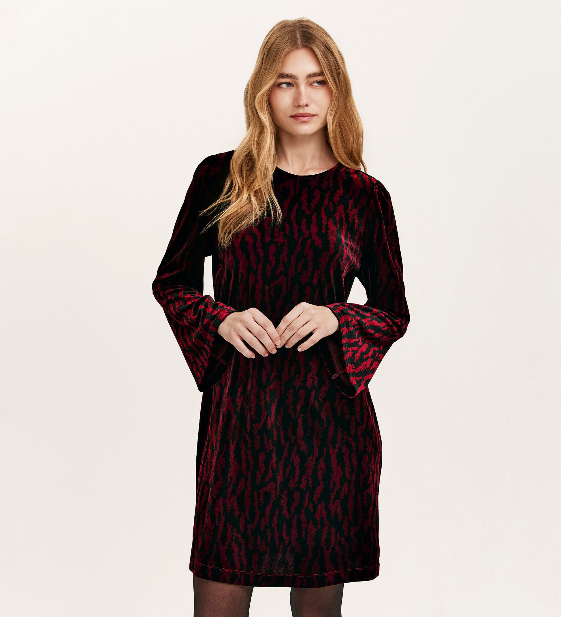 A woman with long blonde hair wears the Hattie Burgundy Zebra Jersey Velvet Short Dress, featuring a red animal print and bell sleeves, perfect for parties, standing against a plain light background.