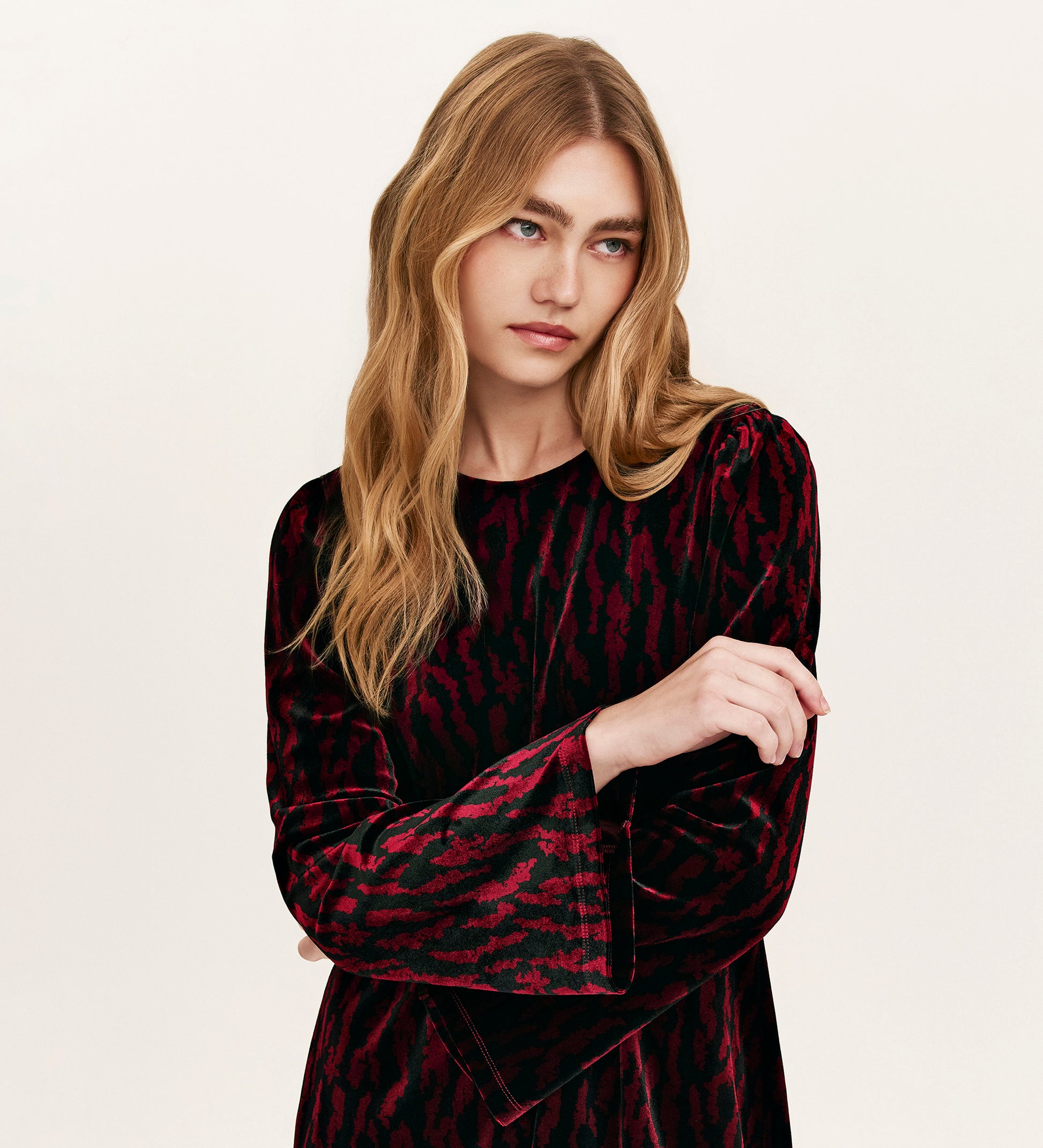 A woman with long, wavy blonde hair wears the Hattie Burgundy Zebra Jersey Velvet Short Dress with long sleeves, arms crossed and a neutral expression—an ideal choice for stylish party dressing against a plain background.