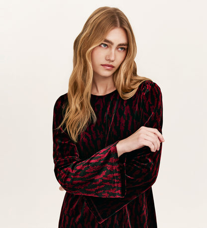 A woman with long, wavy blonde hair wears the Hattie Burgundy Zebra Jersey Velvet Short Dress with long sleeves, arms crossed and a neutral expression—an ideal choice for stylish party dressing against a plain background.