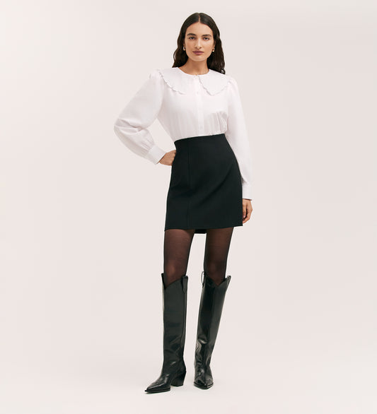 A woman with long dark hair and a neutral expression stands against a plain background wearing a white blouse, sheer black tights, knee-high black boots, and the Helia Black Ponte Jersey Midi Skirt.